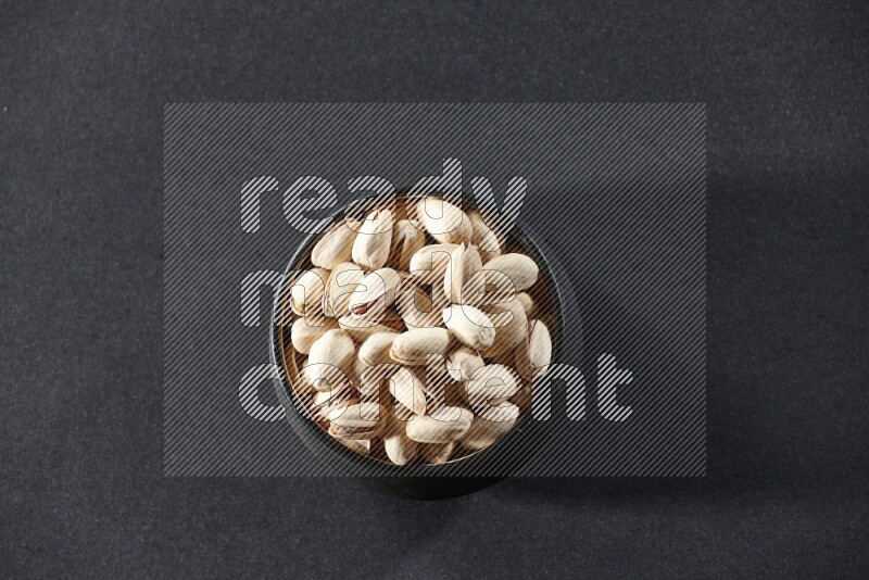 A black pottery bowl full of pistachios on a black background in different angles
