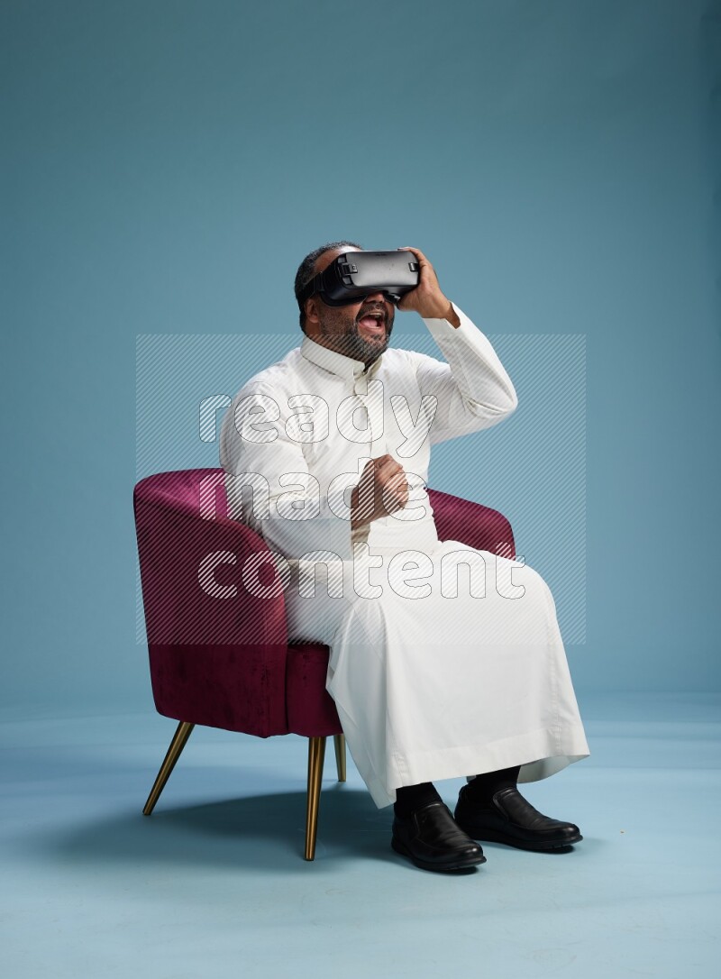 Saudi Man without shimag sitting on chair  gaming with VR on blue background