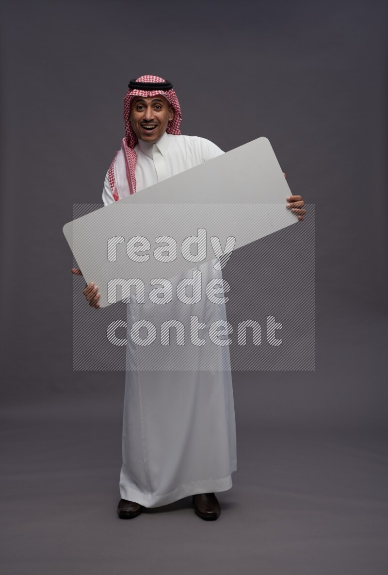 Saudi man wearing thob and shomag standing holding board on gray background