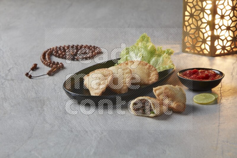 Three fried sambosas in an oval shaped black plate and a red sauce in a black round ramekin with a brown misbaha and a golden lantern on the side on a gray background