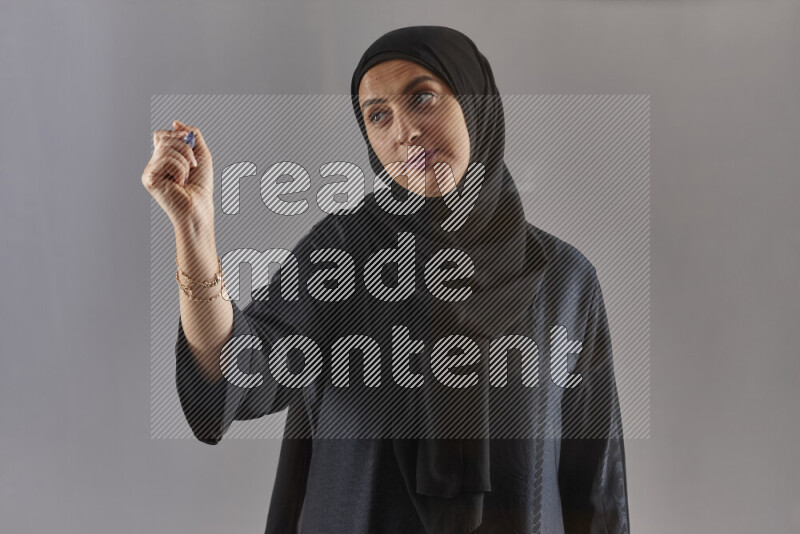 Woman in black abaya and hijab holding a marker pen to write on a transparent board with different reactions and expressions on grey background