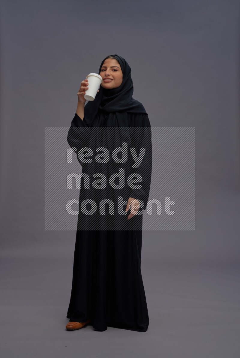 Saudi woman wearing Abaya standing holding paper cup on gray background
