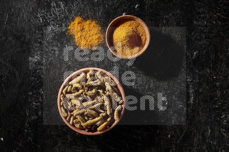 2 wooden bowls, one full of turmeric powder and the other full of dried turmeric whole fingers on a textured black flooring