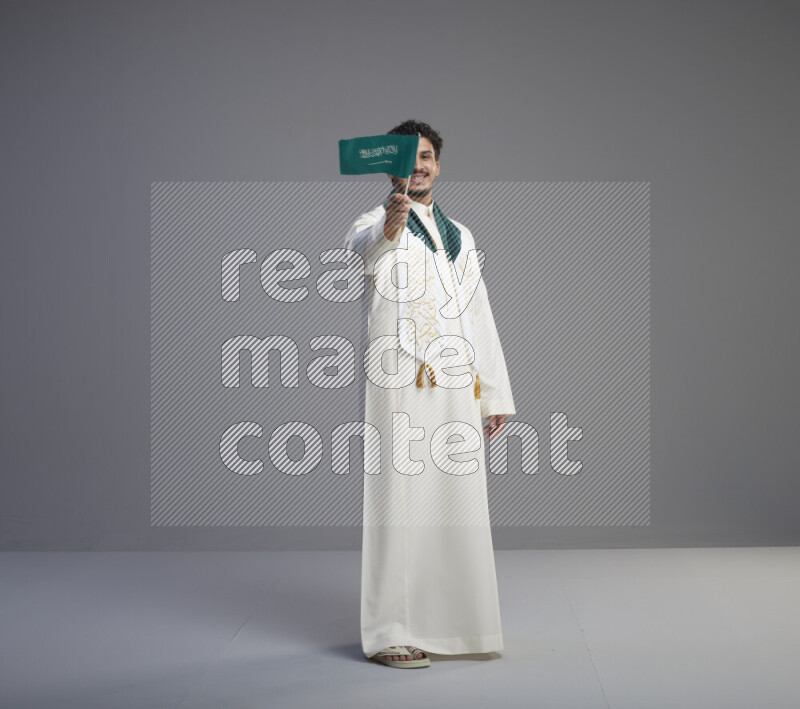 A Saudi man standing wearing thob and saudi flag scarf and holding small saudi flag on gray background