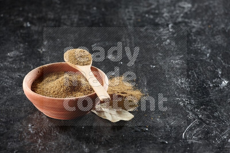 A wooden bowl and spoon full of cumin powder on a textured black flooring