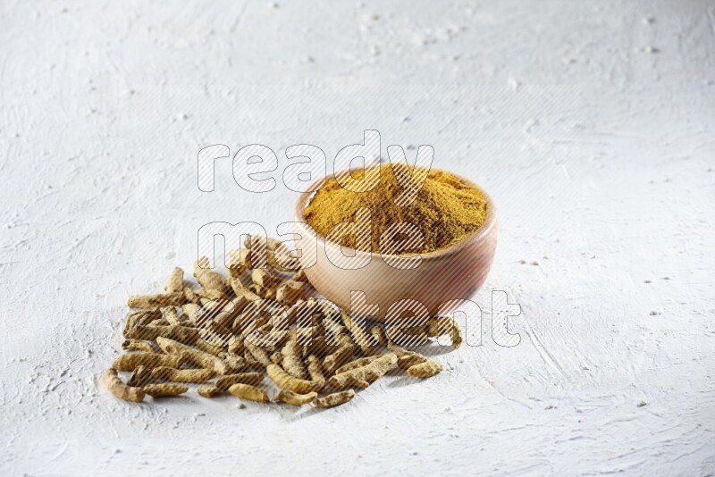 A wooden bowl full of turmeric powder and dried whole fingers beside it on a textured white flooring