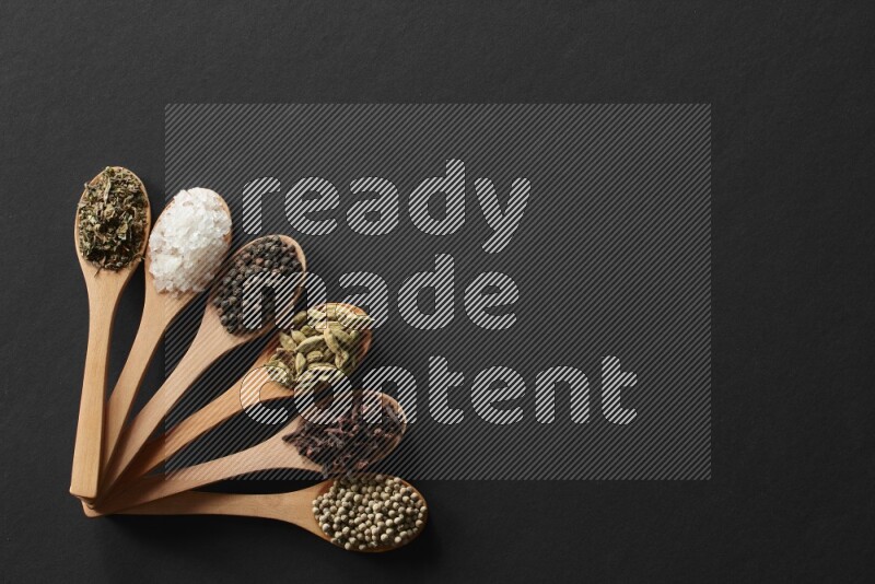 الفلفل الأبيض والقرنفل والهيل والملح والفلفل الأسود وريحان مجفف في معالق خشبية علي خلفية سوداء