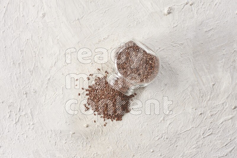 A glass spice jar full of flax seeds flipped and seeds spread out on a textured white flooring