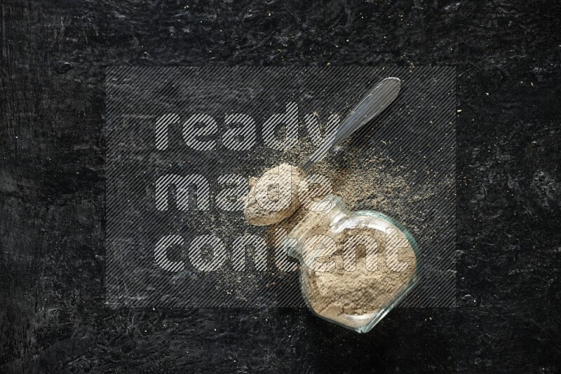 وعاء زجاجي للتوابل مقلوب وملعقة معدنية ممتلئان ببودرة الهيل والبودرة تناثرت منه على خلفية سوداء