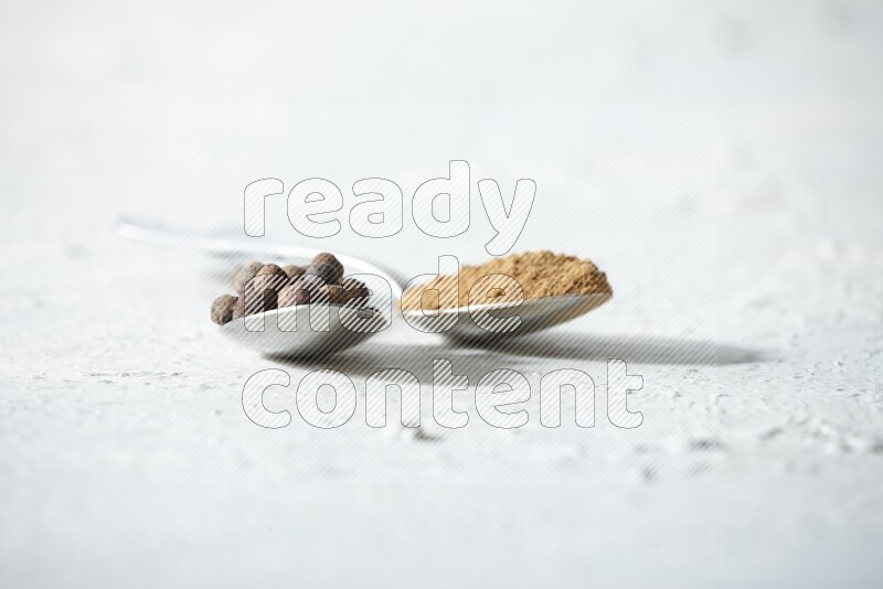 2 metal spoons full of allspice powder and whole balls on a textured white flooring