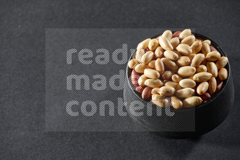 A black pottery bowl full of peeled peanuts on a black background in different angles