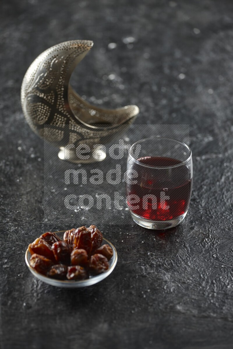 A silver lantern with different drinks, dates, nuts, prayer beads and quran on textured black background