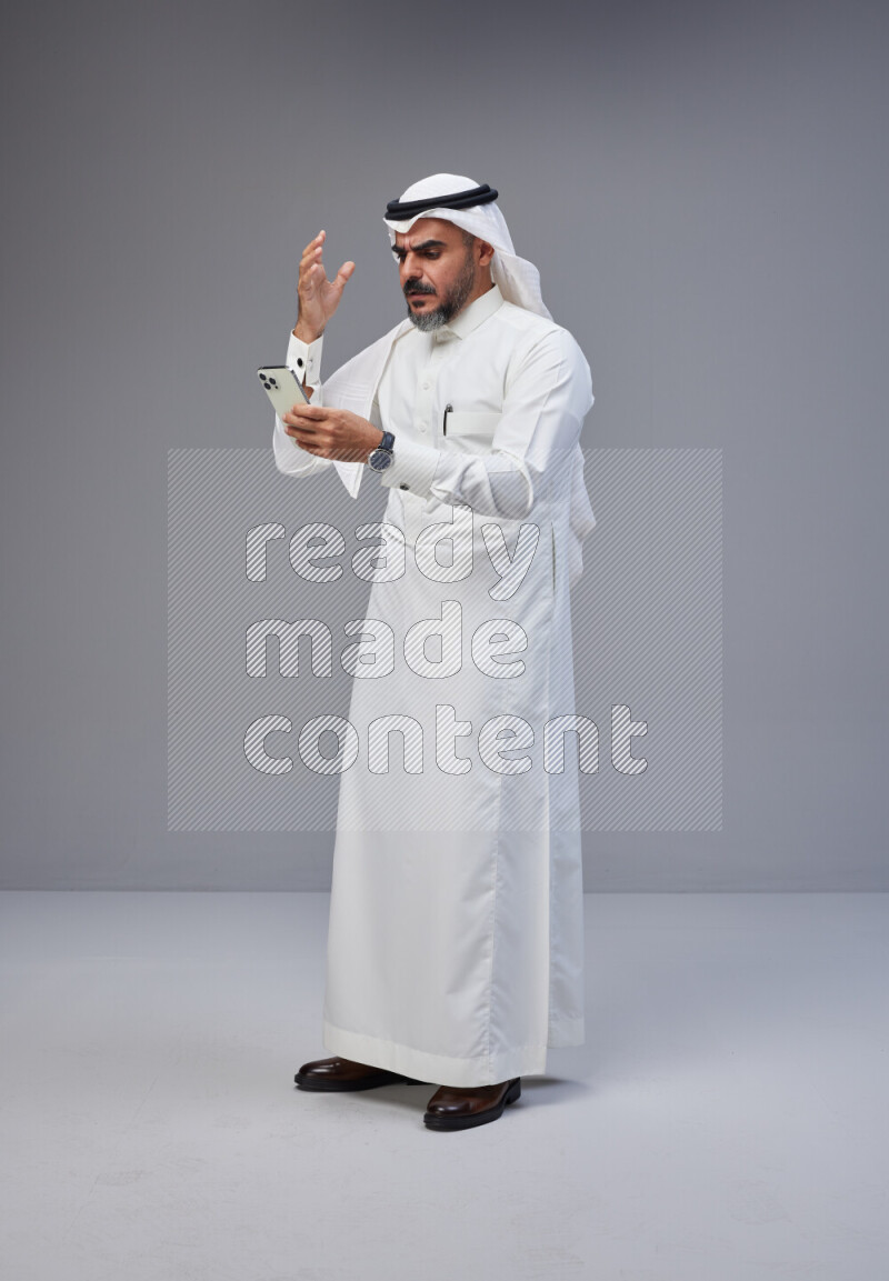 Saudi man Wearing Thob and white Shomag standing texting on phone on Gray background