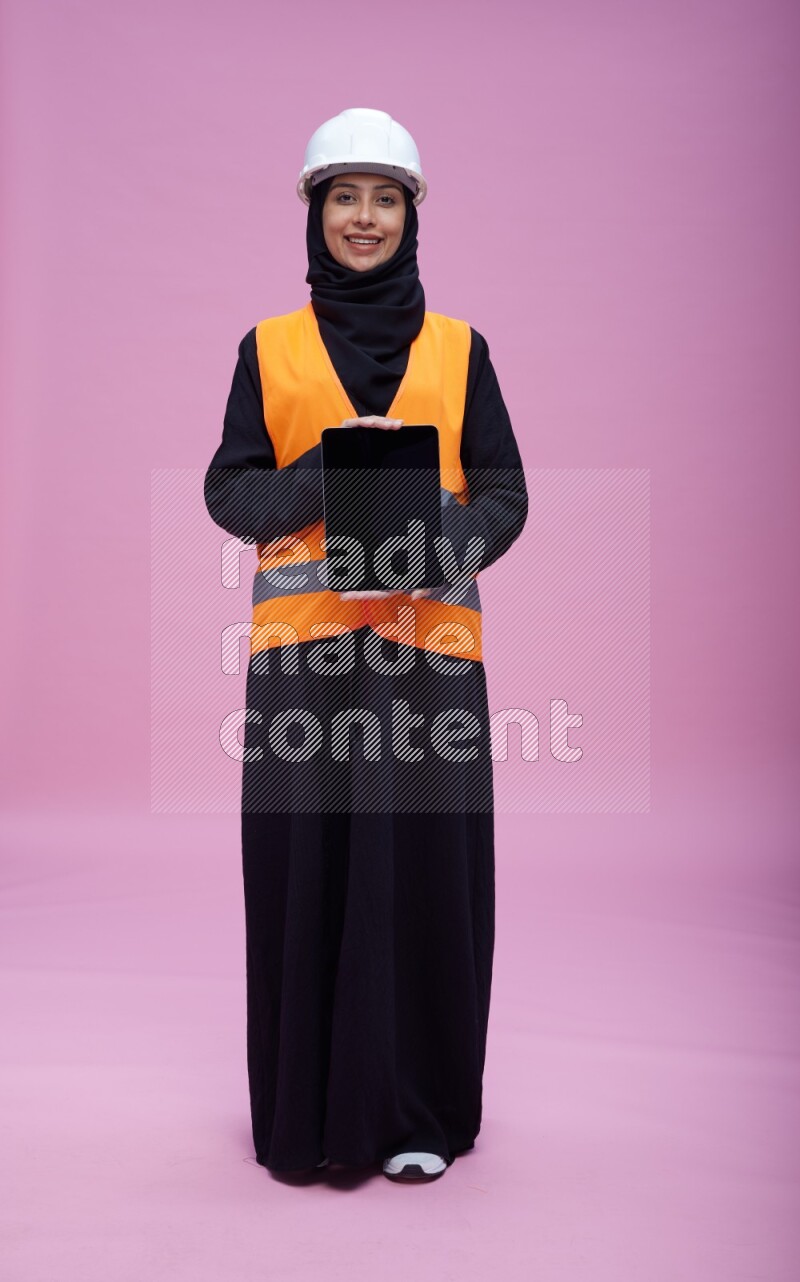 Saudi woman wearing Abaya with engineer vest and helmet standing showing tablet to camera on pink background