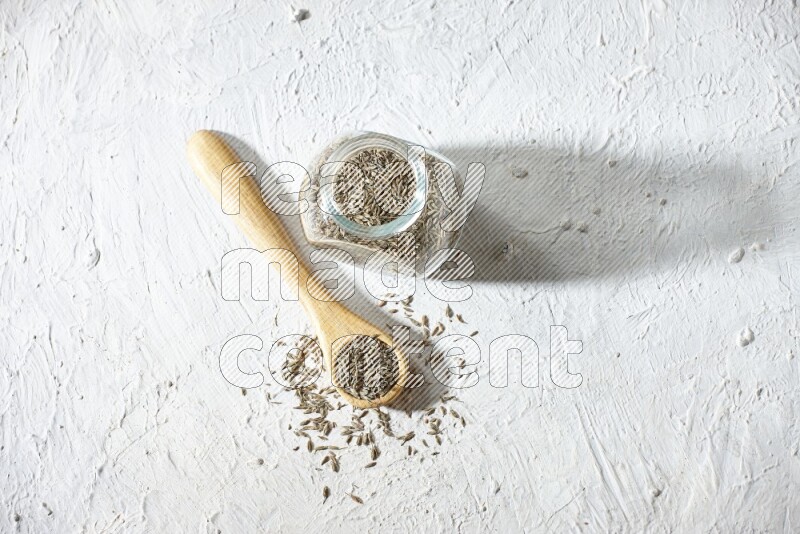 A glass spice jar and wooden spoon full of cumin seeds on textured white flooring
