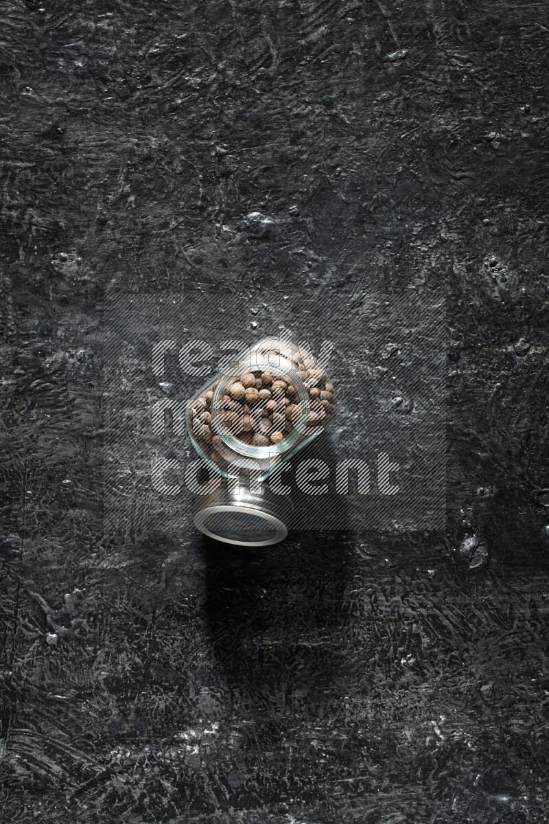 A glass spice jar full of allspice whole balls on a textured black flooring