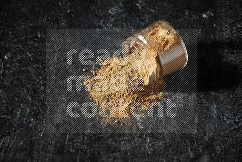 A flipped glass jar full of allspice powder and powder spilled out of it on a textured black flooring