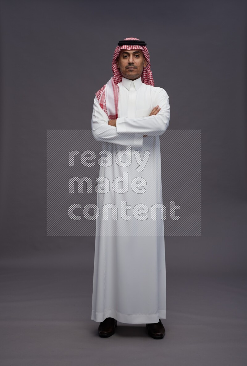 Saudi man wearing thob and shomag standing with crossed arms on gray background