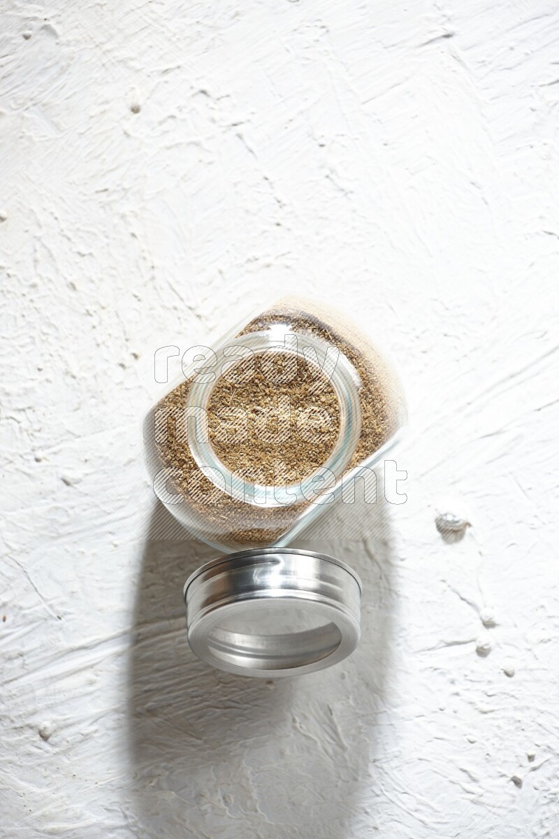 A glass spice jar full of cumin powder on textured white flooring