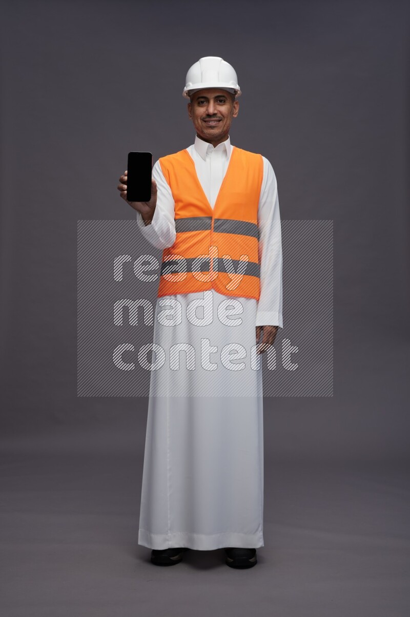 Saudi man wearing thob with engineer vest standing showing phone to camera on gray background