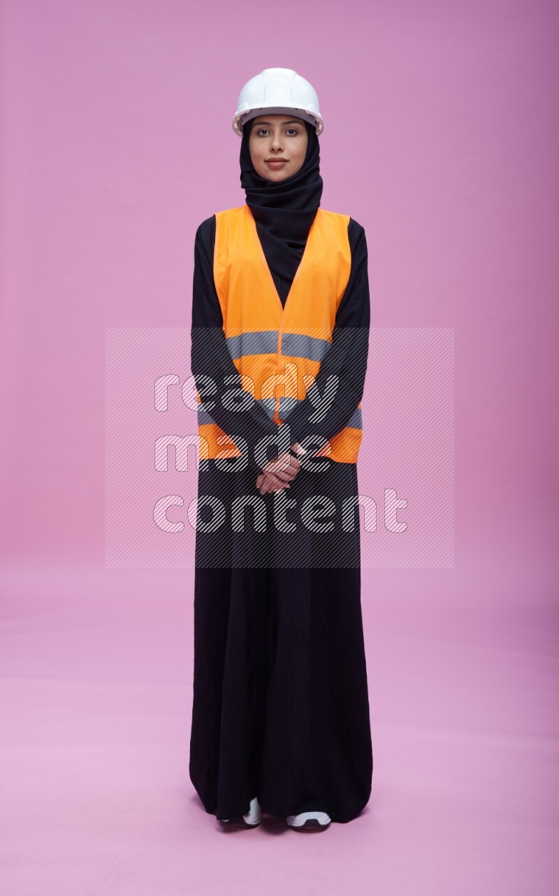 Saudi woman wearing Abaya with engineer vest and helmet standing interacting with the camera on pink background
