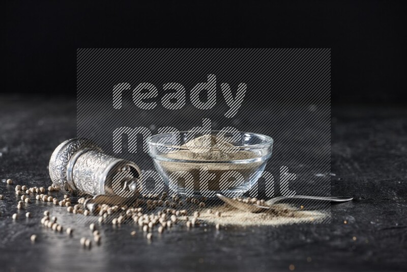 A glass bowl full of white pepper powder with pepper beads, a metal grinder and a metal spoon on textured black flooring