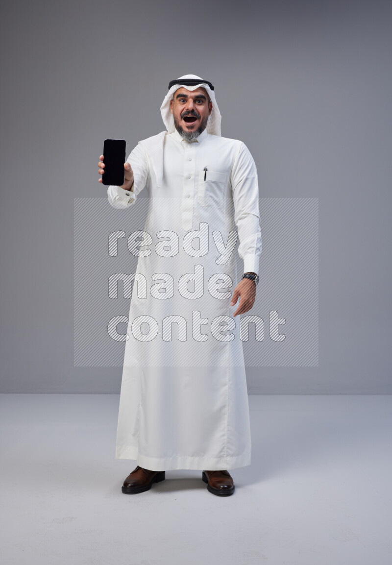 Saudi man Wearing Thob and white Shomag standing showing phone to camera on Gray background