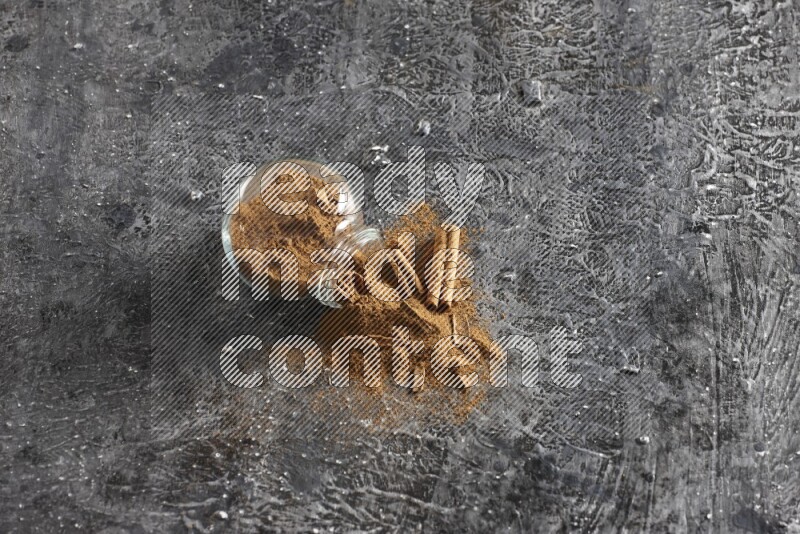 Flipped glass herbs jar full of cinnamon powder with cracked cinnamon sticks on a textured black background