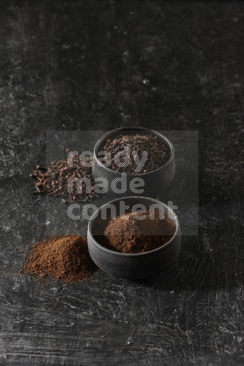2 Black pottery bowls full of cloves and the other full of cloves powder on textured black flooring