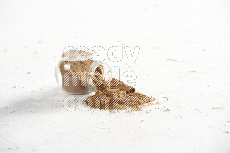 Flipped glass jar full of cinnamon powder with some pieces of cinnamon sticks on a textured white background