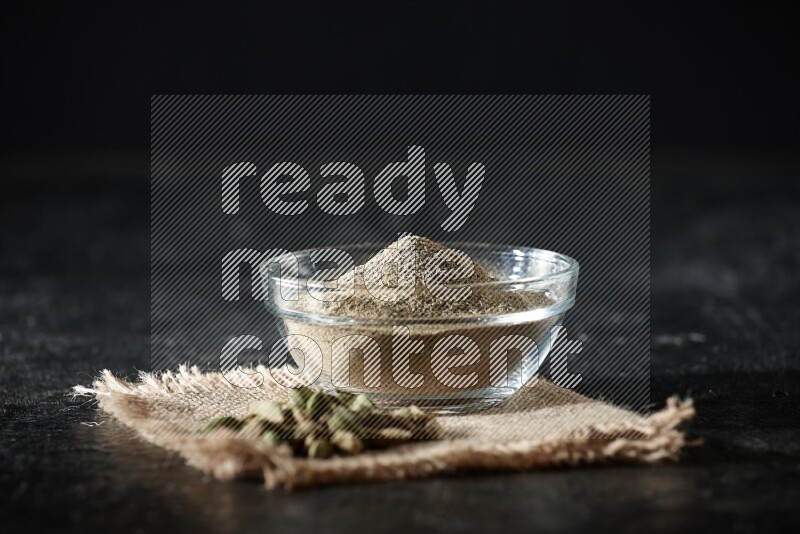 A glass bowl full of cardamom powder with cardamom seeds on a burlap piece on textured black flooring