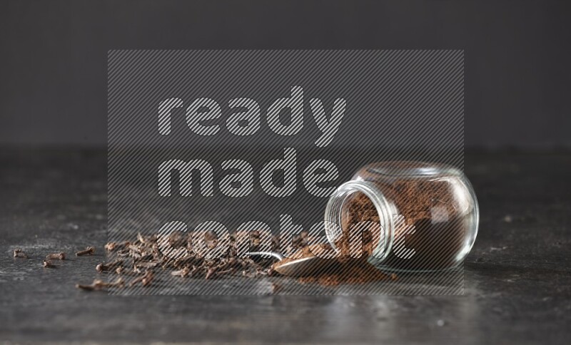A flipped glass spice jar and a metal spoon full of cloves powder with cloves spread on textured black flooring