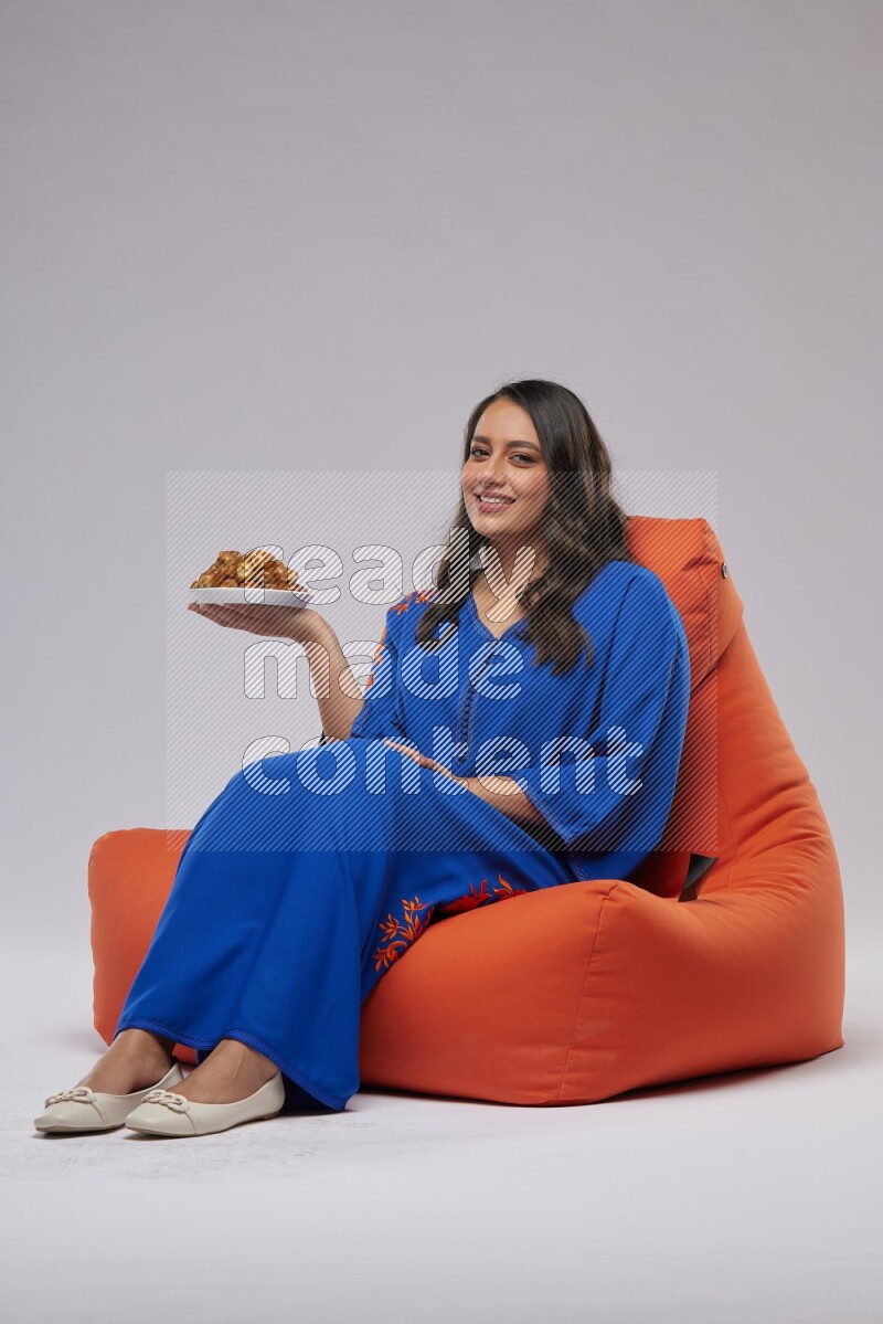 A Woman sitting on an orange beanbag wearing Jalabeya holding a plate of dates