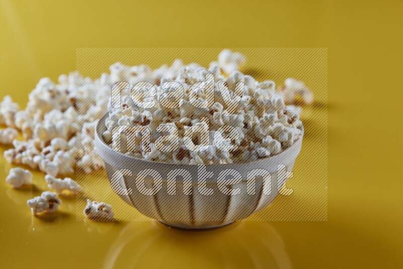 A white pottery bowl full of popcorn with popcorn beside it on a yellow background in different angles