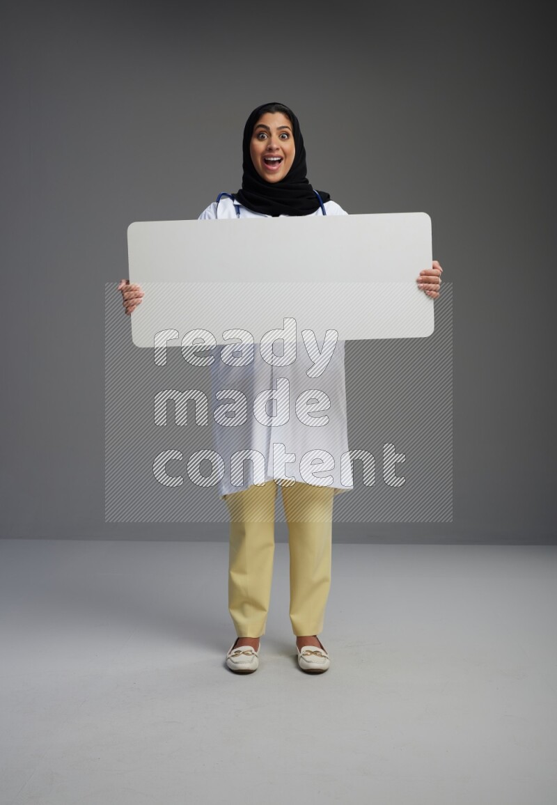 Saudi woman wearing lab coat with stethoscope standing holding board on Gray background