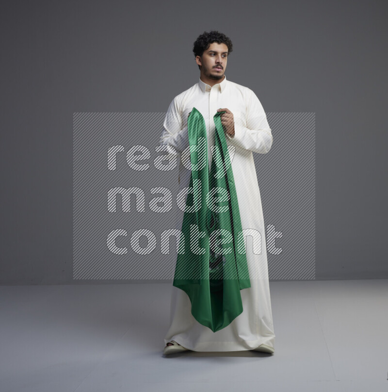 A Saudi man standing wearing thob holding big Saudi flag on gray background