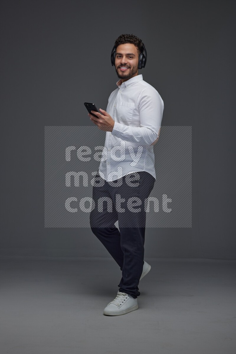 A man wearing smart casual and using his phone and headphone eye level on a gray background