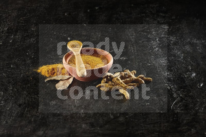 A wooden bowl and a wooden spoon full of turmeric powder with dried turmeric fingers on textured black flooring