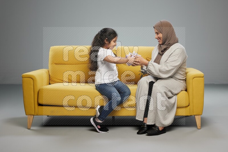 A girl sitting on a yellow sofa and giving a gift to her mother on gray background