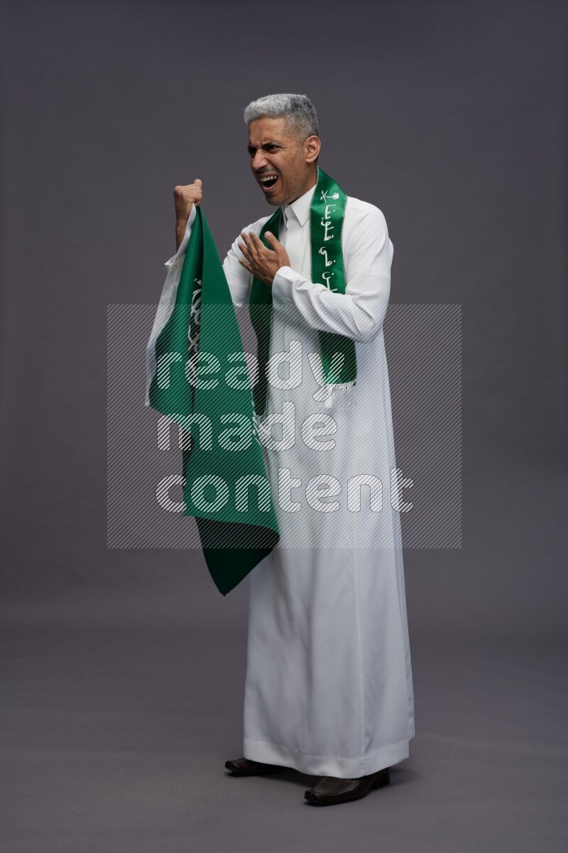 Saudi man wearing thob standing holding Saudi flag on gray background