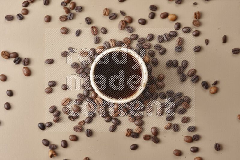 A beige pottery cup of coffee surrounded by roasted coffee beans on beige background