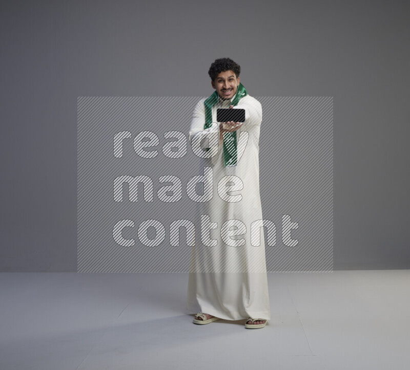 A Saudi man standing wearing thob and Saudi flag scarf showing phone to camera on gray background