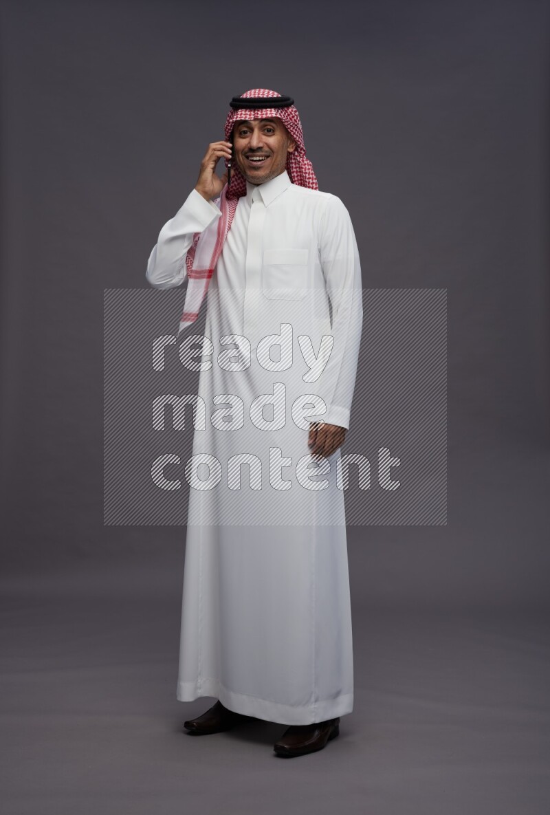 Saudi man wearing thob and shomag standing talking on phone on gray background