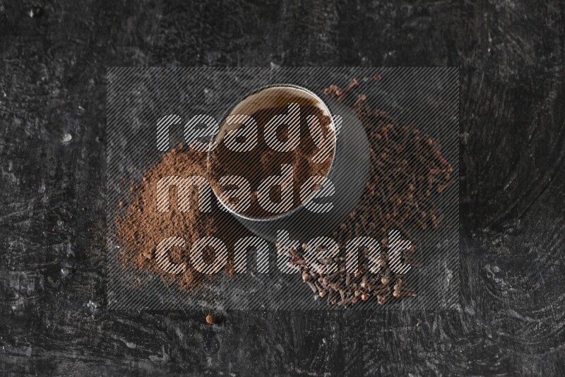 وعاء فخاري أسود ممتلئ ببودرة القرنفل والبودرة تناثرت منه مع الحبوب على خلفية سوداء