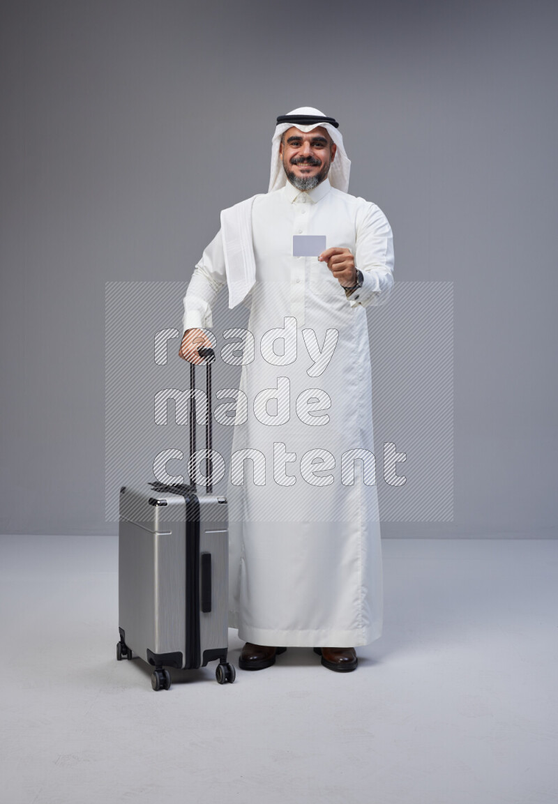 Saudi man wearing Thob and white Shomag standing holding Travel bag and ATM card on Gray background