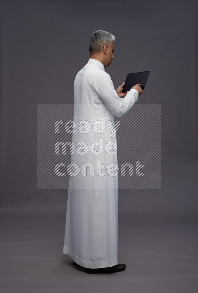 Saudi man wearing thob standing working on tablet on gray background