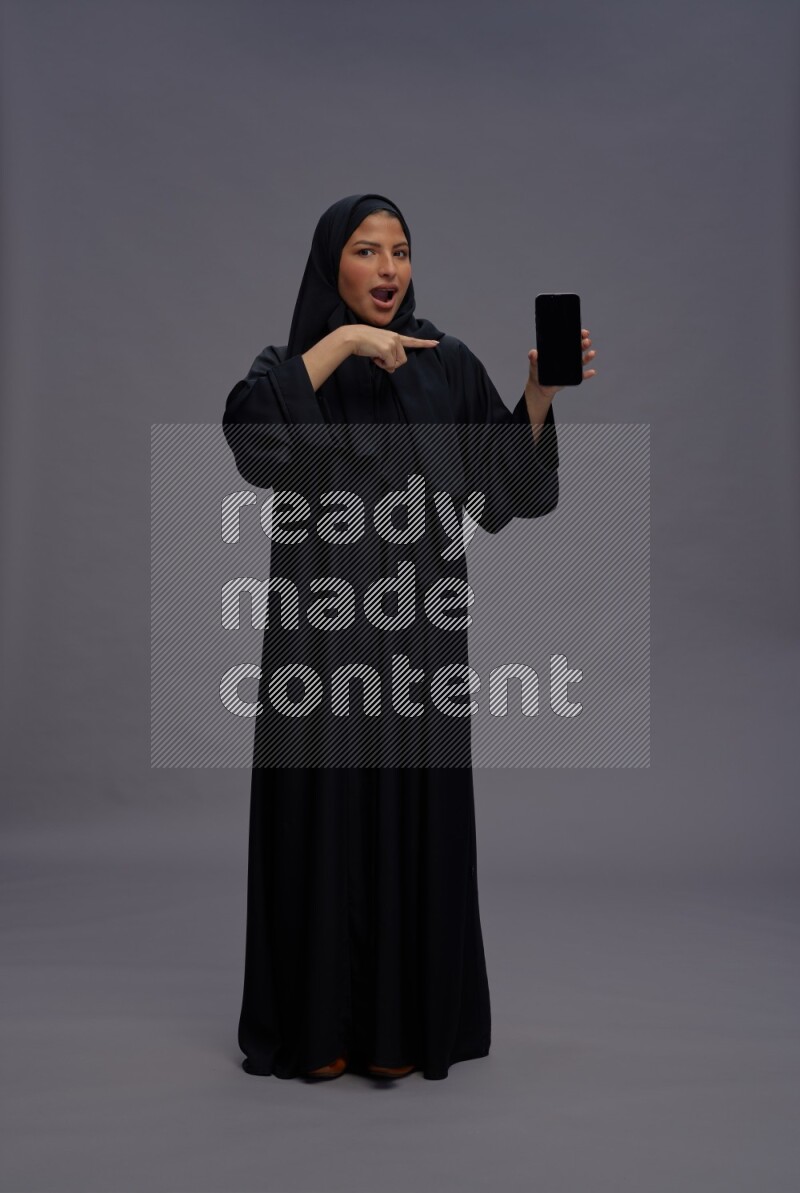 Saudi woman wearing Abaya standing showing phone to camera on gray background