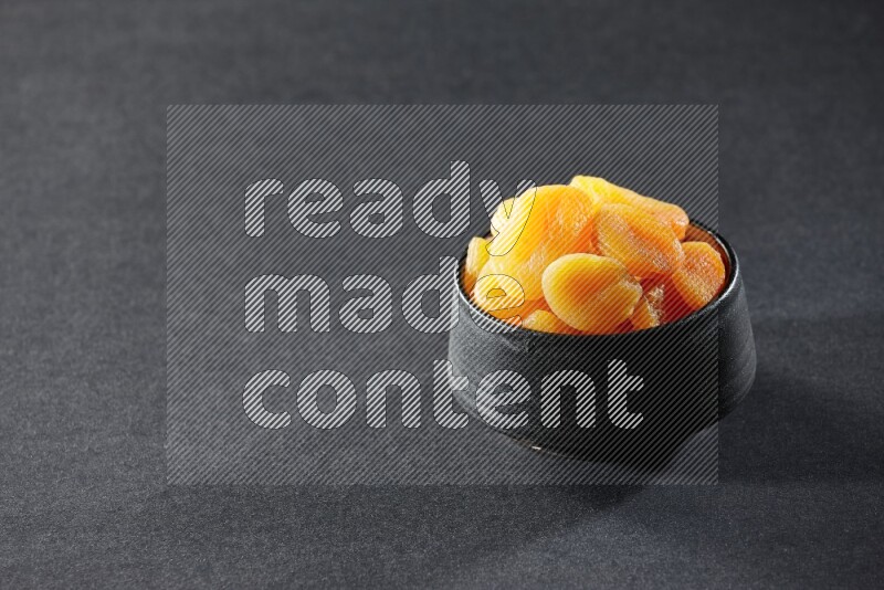 A black pottery bowl full of dried apricots on a black background in different angles