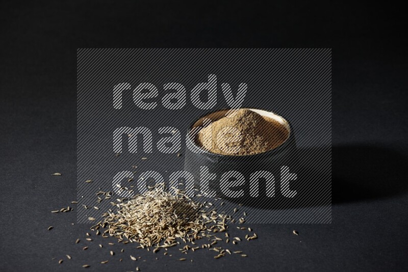 A black pottery bowl full of cumin powder and spreaded cumin seeds on a black flooring