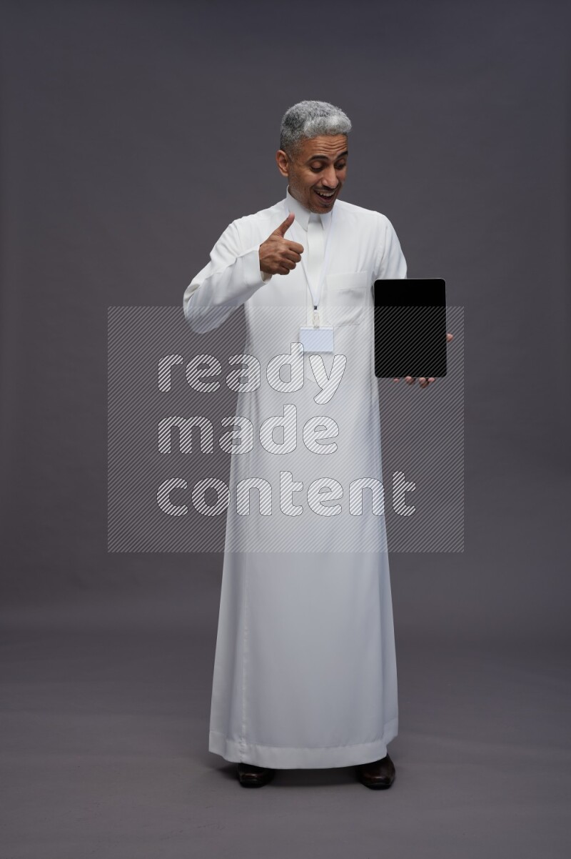 Saudi man wearing thob with neck strap employee badge standing showing tablet to camera on gray background